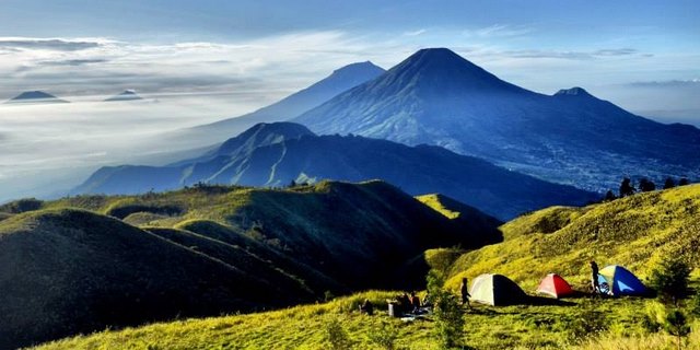 Keindahan Perkemahan Puncak Gunung Prau: Pengalaman Trip Gunung Prau yang Tak Terlupakan Pemandangan perkemahan di puncak Gunung Prau dengan tenda-tenda berwarna di padang rumput hijau dan latar belakang pegunungan biru yang diselimuti kabut saat matahari terbit, sempurna untuk trip Gunung Prau.